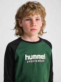 Barn med lyst krøllet hår og fregner iført en grøn og sort langærmet trøje med teksten "hummel SPORTSWEAR" trykt på fronten.