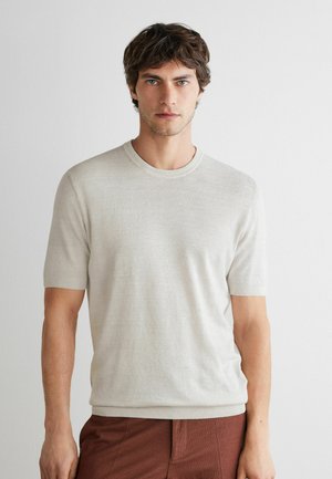 Ung mand med bølget brunt hår iført en lysebeige kortærmet striktrøje og brune bukser mod en ensfarvet hvid baggrund.