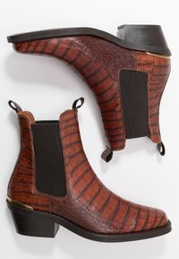Bottines en cuir crocodile brun avec des panneaux latéraux en élastique et des talons courts, présentées en angle pour mettre en valeur les deux côtés sur un fond blanc.