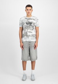 Ανοιχτό γκρι παντελόνι camo T-shirt με μαύρο λογότυπο, σε συνδυασμό με γκρι τζιν σορτς. Το μοντέλο φοράει ανοιχτό γκρι αθλητικά παπούτσια και λευκές κάλτσες. Απλή, καθημερινή εμφάνιση.