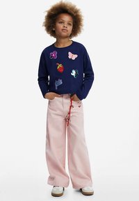 Marineblauw shirt met lange mouwen en kleurrijke patches; lichtroze jeans met wijde pijpen en rode accenten; crème sneakers met een dikke zool.