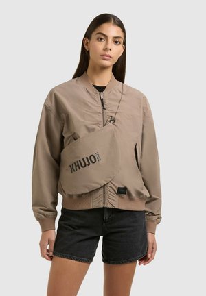 Nő, aki taupe színű bomber dzsekit és keresztpántos táskát visel "KHUJO 2001" felirattal feketén, fekete farmer rövidnadrággal, egyszínű háttér előtt.