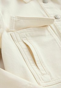 Gros plan sur une poche de veste blanc cassé avec un rabat, des coutures visibles et deux boutons sur le coin supérieur droit.
