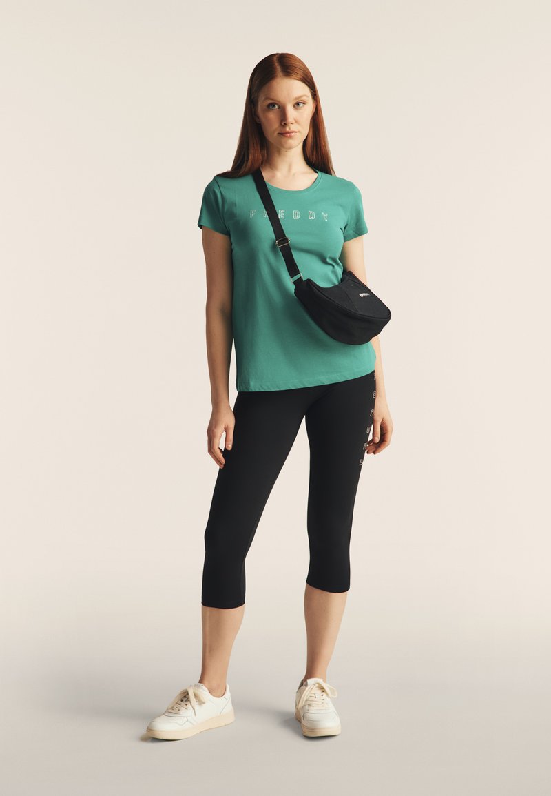 T-shirt di cotone verde con stampa "FREDDY", leggings neri e scarpe da ginnastica bianche. Gli accessori includono una borsa a vita nera. Texture lisce in tutto l'outfit.