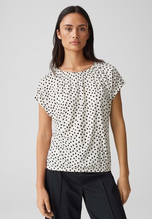 Haut court à manches courtes blanc avec des pois noirs, encolure froncée et ourlet élastique. Associé à un pantalon sombre.