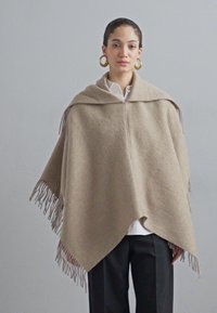 Poncho en laine beige avec une capuche, présentant des franges le long de l'ourlet et une forme triangulaire. Porté par-dessus une chemise blanche et un pantalon noir.
