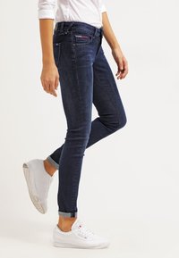 Vaqueros ajustados de mezclilla azul oscuro, diseño ceñido, bolsillos delanteros y traseros, con los puños enrollados, combinados con zapatillas blancas para un contraste.