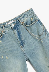 Jeans de mezclilla azul claro con detalles desgastados y un accesorio de cadena plateada a lo largo del lado. Diseñados con un cierre de botón y cremallera.
