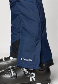 Tengerészeti sínadrág, Columbia logóval az alján, részletes varrással és szűkített fazonnal, fekete sícipőkkel, ezüst részletekkel párosítva.