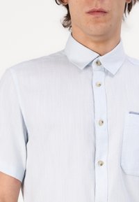 Light blue short-sleeve shirt made of textured fabric. Features a button-down collar, front buttons, and a single chest pocket.
