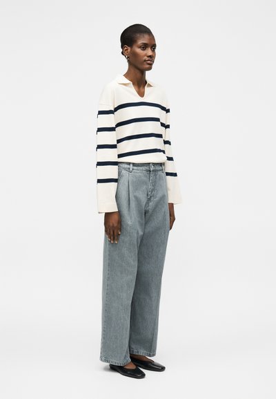 Νέα γυναίκα που στέκεται, φοράει λευκό μπλουζάκι με μακριά μανίκια και μαύρες οριζόντιες ρίγες, άνετο γκρι τζιν και μαύρα επίπεδα παπούτσια.