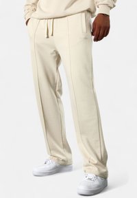 Creamfarbene Sweatpants aus weichem Baumwollmaterial, mit geradem Schnitt, seitlichen Taschen und kontrastierenden Nähten. Getragen mit weißen Turnschuhen.