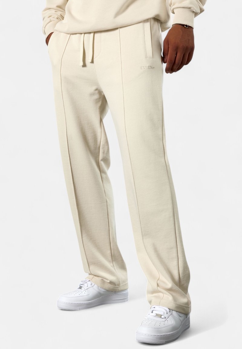 Creamfarbene Sweatpants aus weichem Baumwollmaterial, mit geradem Schnitt, seitlichen Taschen und kontrastierenden Nähten. Getragen mit weißen Turnschuhen.