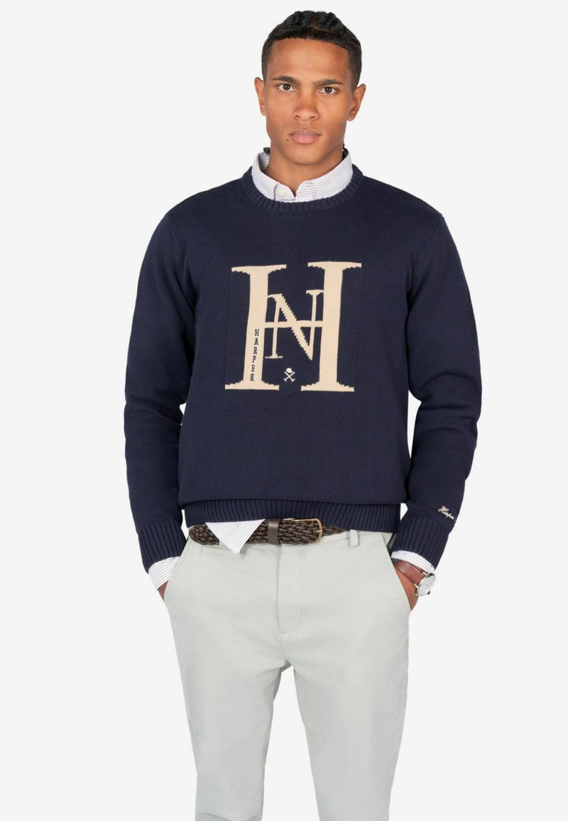 Suéter de punto azul marino con un gran logo beige que presenta las letras "HN" y texto. Puños y dobladillo acanalados. Lleva una camisa de color azul claro con cuello debajo.