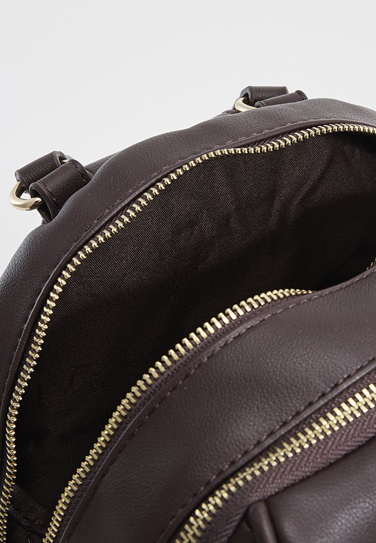 Brauner Leder-Rucksack mit goldenen Reißverschlüssen. Das Innenfutter besteht aus einem dunklen Stoff. Doppelte Reißverschlussfächer sind sichtbar. Verstellbare Träger sind vorhanden.