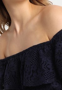 Žena v tmavě modrém krajkovém topu na jedno rameno, který odhaluje klíční kost a rameno, má vlnité hnědé vlasy, které jsou částečně viditelné.