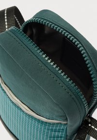 Türkizkék nejlon válltáska csíkozott textúrával, cipzáros rekesszel és állítható pánttal. A belső rész egyszínű anyagbéléssel van ellátva a védelem érdekében.