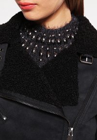 Donna che indossa una giacca di shearling nera sopra un maglione morbido grigio scuro abbellito con perline nere lucide.