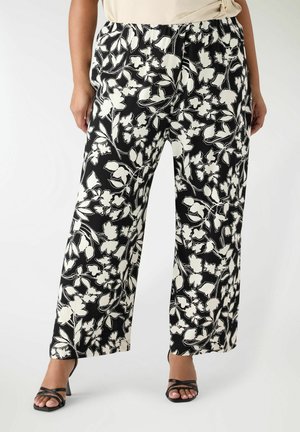 Pantaloni neri larghi con stampa floreale bianca indossati con tacchi neri con cinturini e top beige, mostrati dalla vita fino ai piedi.