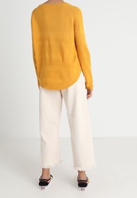 Sweater en tricot moutarde avec une finition texturée, associé à un pantalon large blanc avec des ourlets effilochés et des baskets noires avec des accents colorés.