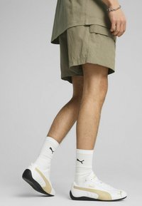 Bijele tenisice s zlatnim detaljima, uparene s bijelim rebrastim čarapama koje imaju crni logo, nosene s maslinasto zelenim cargo kratkim hlačama.