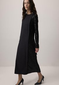 Vestido negro con una manga de lentejuelas, con un panel negro sólido y un detalle de lazo en la cintura. Diseño cosido, estilo de longitud media.