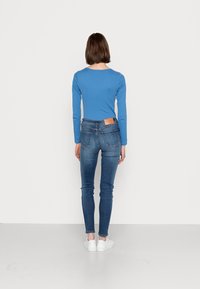 Langarmige blauwe top en slim-fit blauwe jeans, met een bruine leren patch op de achterkant van de taille en witte sneakers. Effen achtergrond.