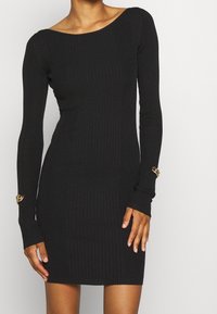 Czarna prążkowana sukienka-sweter z długimi rękawami, szerokim dekoltem i unikalnymi złotymi ozdobami przy nadgarstkach. Dopasowany krój.