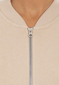 Veste zippée beige avec un col côtelé et une fermeture éclair argentée. Le tissu est lisse, avec une texture légère et des détails de couture minimaux.