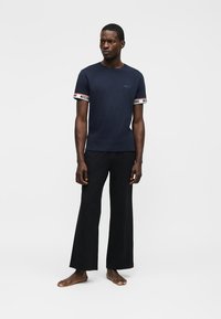 Homme debout pieds nus portant un t-shirt bleu marine avec le logo Moschino sur les manches et un pantalon noir à jambes larges sur fond blanc.
