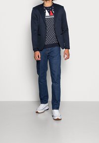 Blazer bleu marine sur un pull noir à motifs d'arbres rouges et blancs, associé à un jean bleu et des baskets blanches à semelles marron.