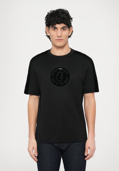 T-shirt en coton noir à manches courtes, avec un accent velouté sur les épaules et un logo circulaire embossé représentant un cheval.