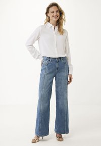 Camicia bianca leggera con bottoni abbinata a jeans blu a gamba larga con vita alta, dotati di tasche anteriori e lavaggio sbiadito, completata da sandali con tacco dorato.