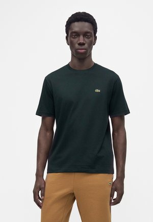 Jeune homme debout face à l'avant, portant un t-shirt vert foncé et un pantalon marron clair avec de petits logos brodés sur les deux vêtements.