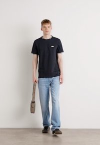 Juodas marškinėliai su kontrastingu apykakle, šviesiai mėlyni džinsai ir rudas peties krepšys. Modelis stovi prieš paprastą foną.