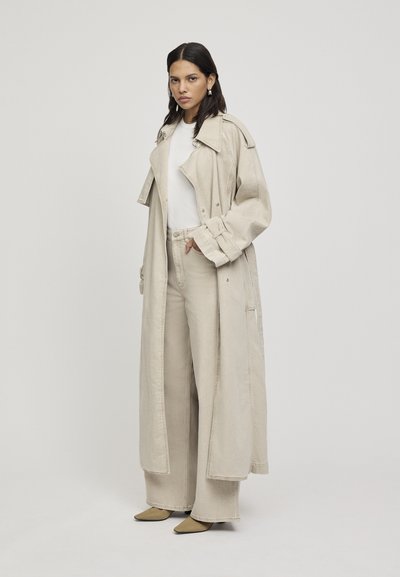 Cappotto trench beige lungo sopra una maglietta bianca, abbinato a pantaloni larghi coordinati e scarpe con punta. Tessuto liscio con dettagli minimi.