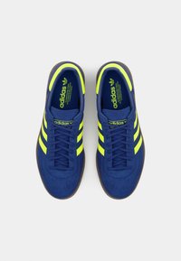 Modré semišové tenisky se žlutými pruhy, s kulatou špičkou, plochými tkaničkami a hnědou gumovou podrážkou. Logo Adidas je viditelné na jazyku.