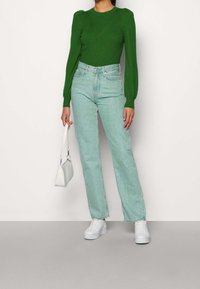 Zielony sweter z dzianiny o puffowanych rękawach, zestawiony z jasnoniebieskimi spodniami o wysokim stanie. Trzyma małą białą torebkę i nosi białe trampki.