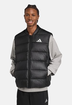 Junger Mann lächelt, trägt eine schwarze gesteppte Weste über einem grauen Sweatshirt und schwarzen Hosen und steht mit den Händen in den Taschen der Weste.