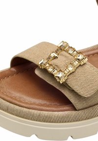 Beige tygsandal med en strukturerad rem, en brun vadderad fotseng och ett guldformat spänne dekorerat med klara rektangulära rhinstenar.