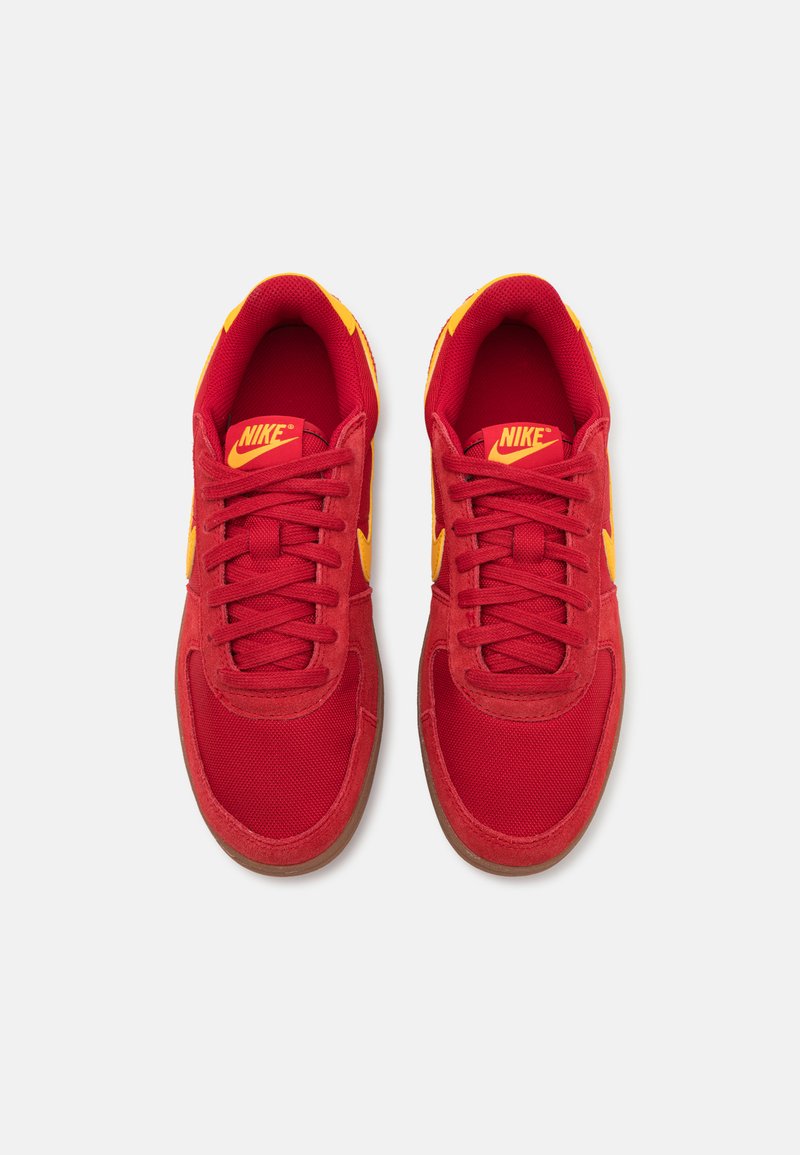 Zapatillas Nike rojas con acentos amarillos, tejido texturizado y cordones planos. El diseño presenta una punta redondeada y una suela de goma.