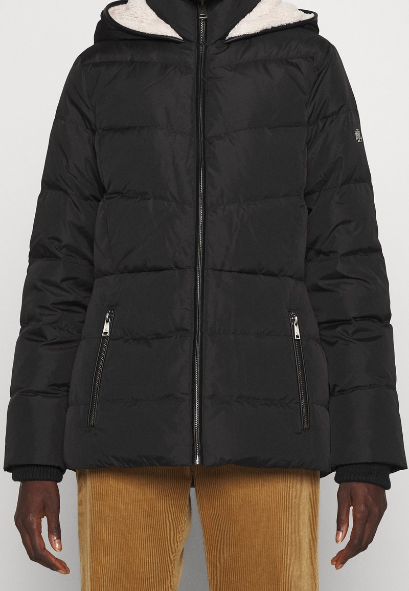 Veste matelassée noire avec motif matelassé, fermeture zippée, deux poches latérales et capuche doublée en matière crème texturée.