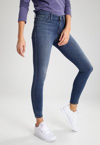 Úzké džíny z tmavě modrého denimu, s středním pasem, designem s pěti kapsami a lehkým vyblednutím. Spárovány s bílými sportovními teniskami.