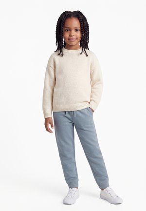 Niño con el cabello trenzado que lleva un suéter crema, pantalones deportivos azul claro y zapatillas blancas, de pie con una mano en el bolsillo.