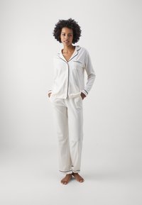 Ljust beige pyjamakläder i mjukt tyg, med svarta detaljer, knäppt topp med ficka och löst sittande byxor.