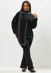 Mustekas musta neule, jossa on valkoista ompelua ja kiinnitetty huivi; yhdistetty mustiin leveisiin housuihin ja mustiin kärkitossuihin.