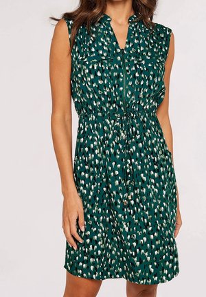 Sleeveless green dress with black, white, and olive abstract spots, front zipper, chest pockets, and waist tie on a standing woman.