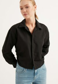 Schwarzes Hemd mit Knöpfen, das einen Kragen, lange Ärmel und eine lockere Passform aufweist, kombiniert mit hellblauen Jeans aus Denim. Glatte Stofftextur.