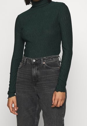 Maglia verde a maniche lunghe realizzata in tessuto testurizzato con colletto alto, abbinata a jeans in denim neri a vita alta con design classico a cinque tasche.