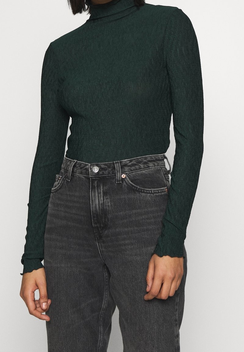 Grünes langärmliges Oberteil aus strukturiertem Stoff mit hohem Kragen, kombiniert mit hoch taillierten schwarzen Jeans aus Denim mit klassischem Fünf-Taschen-Design.